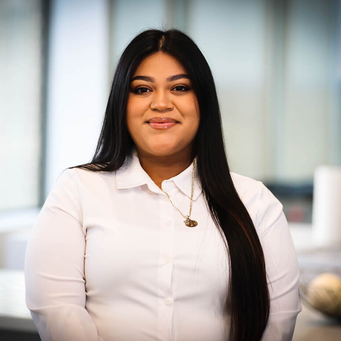 A smiling woman with long dark hair, wearing a white shirt and a necklace, posing in front of an office setting.