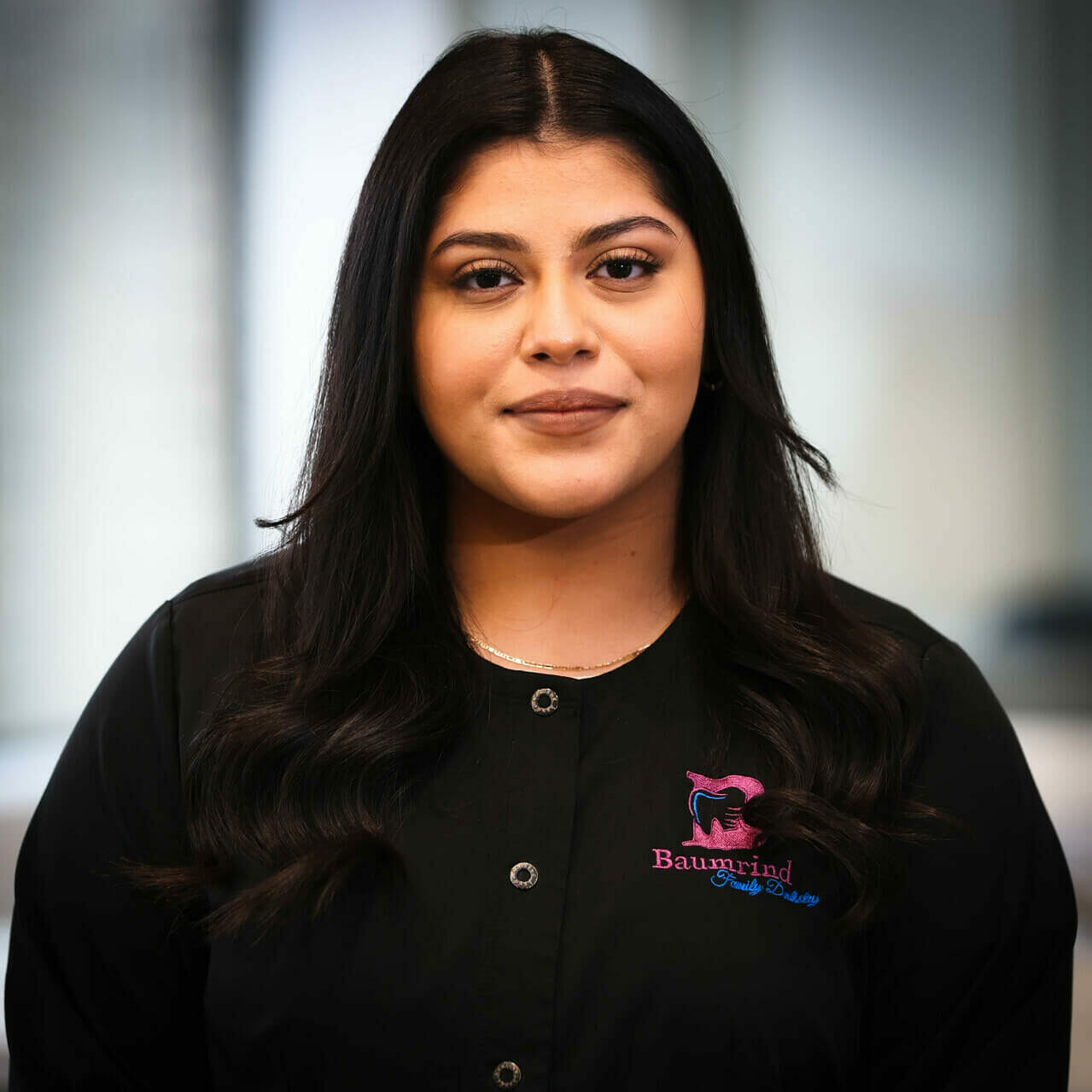 The image is a photograph of a woman standing in front of a background with text. She appears to be wearing a black jacket and has dark hair. Her facial expression is neutral, and she is looking directly at the camera.
