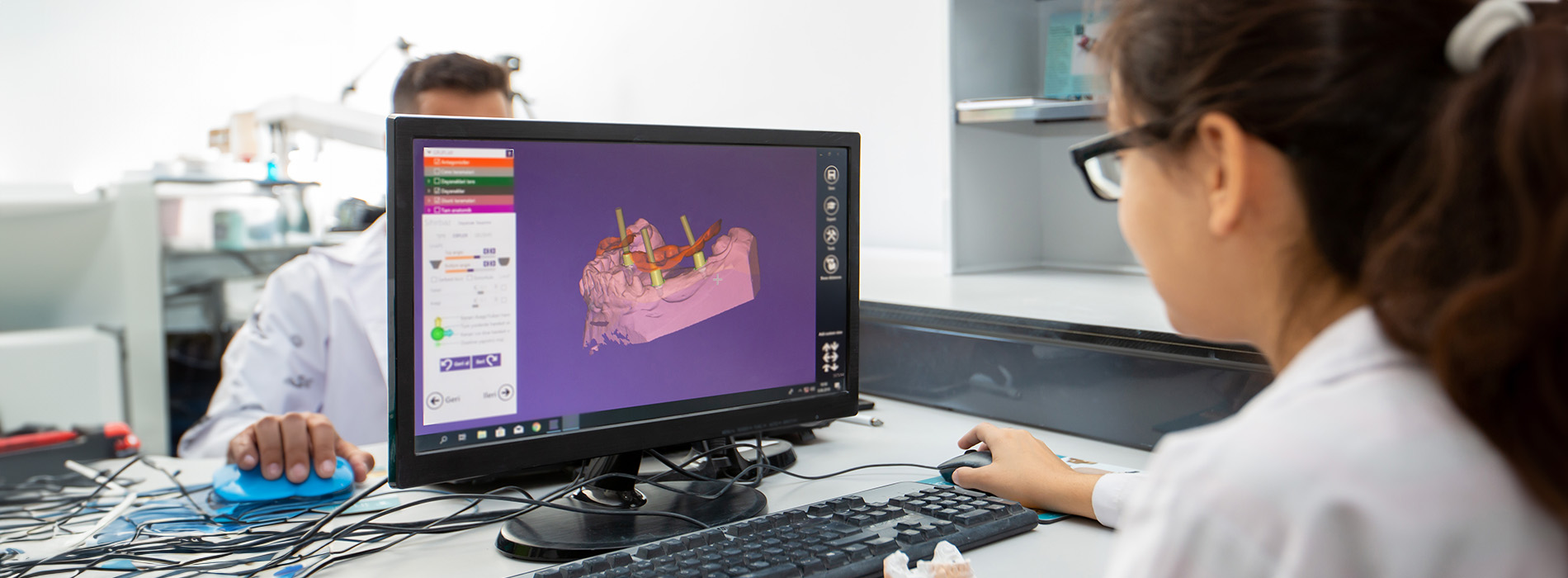 A person sitting at a computer with a monitor displaying a 3D model, surrounded by a lab setting with scientific equipment in the background.