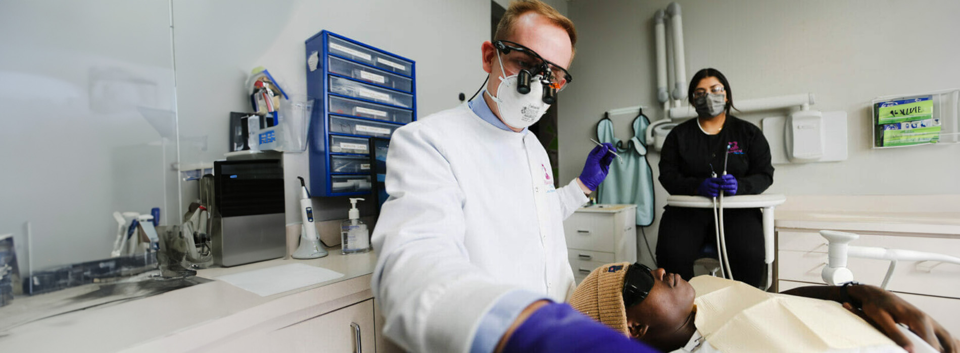 A person in a lab coat is attending to another person who appears to be receiving medical treatment, with various pieces of equipment visible in the background.