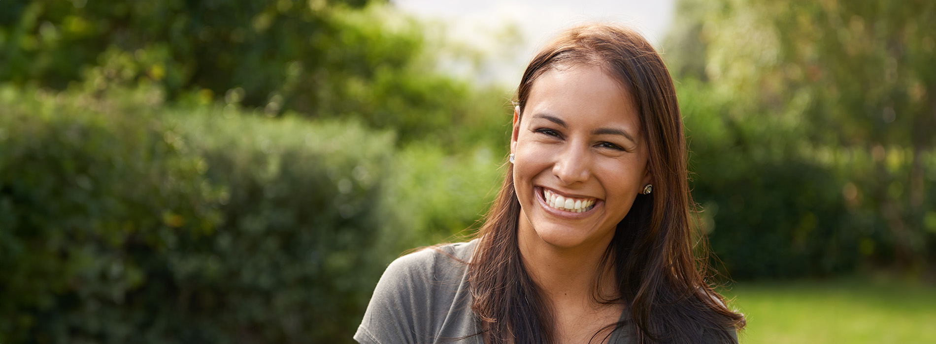 A smiling woman in a park setting, with her head turned to the side, looking at something off-camera.