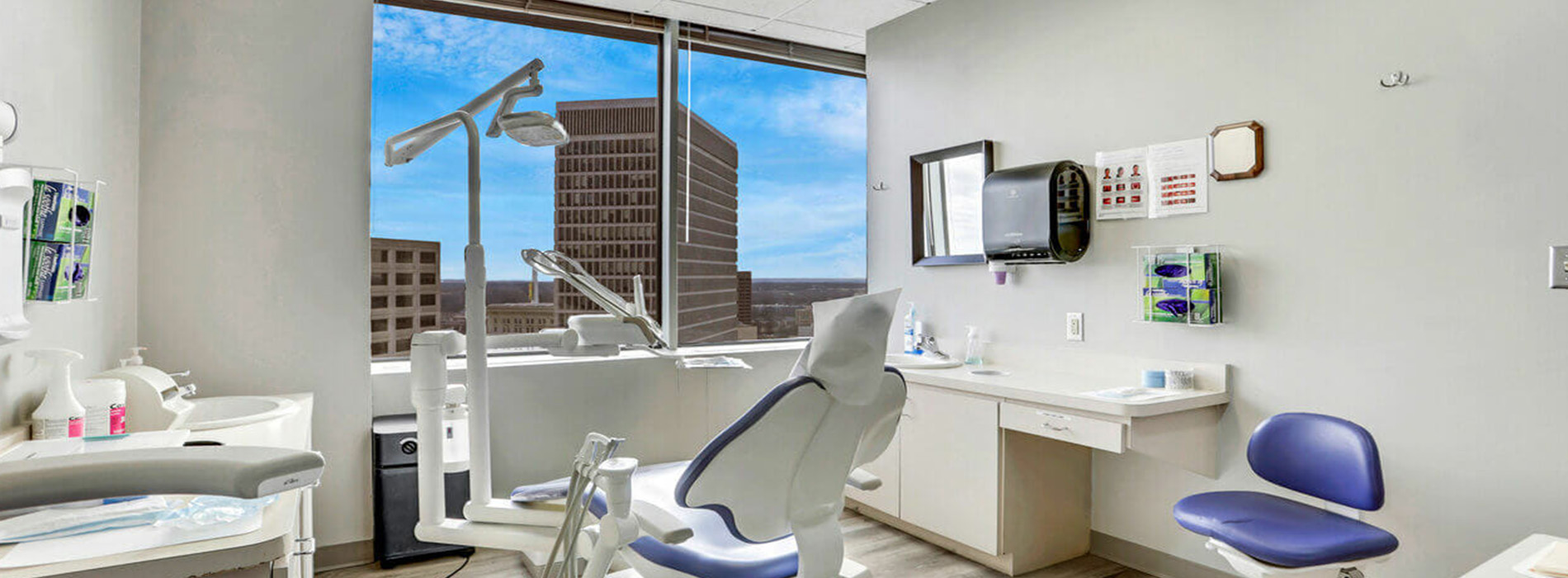 The image shows a modern dental office interior with a large window offering a view of the city skyline, a dental chair in the foreground, and various dental equipment visible.