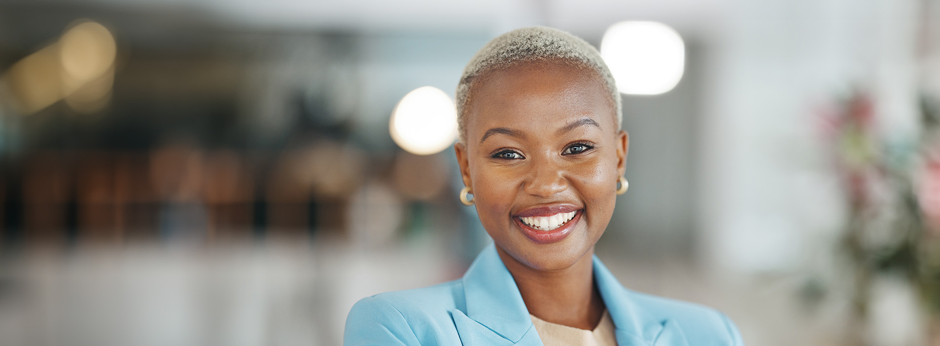 A smiling woman with short hair, wearing a blue blazer and standing in an indoor setting with blurred background.