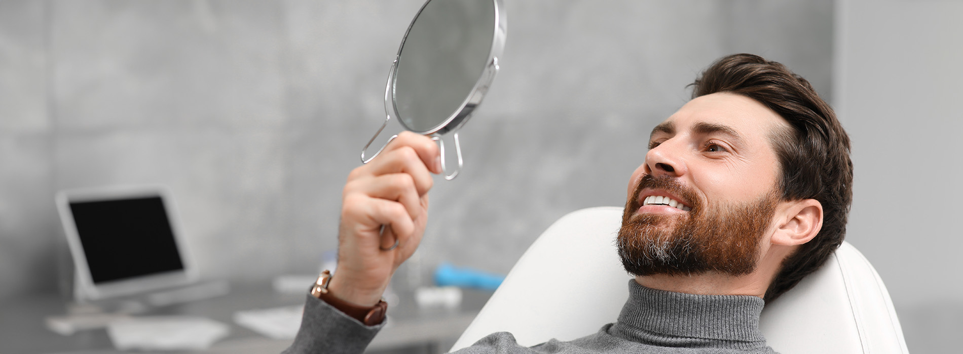 A man in a professional setting looking at his reflection in a mirror while holding a smartphone.