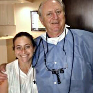 An elderly man in a blue surgical gown and a woman are posing for the camera, with the man wearing glasses and holding a dental mirror.