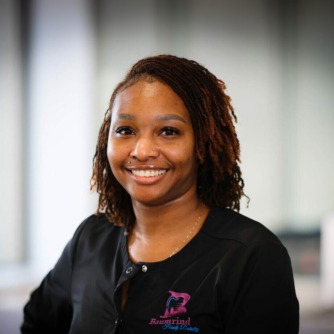The image is a portrait of a smiling woman wearing a black shirt, posing against a blurred background.