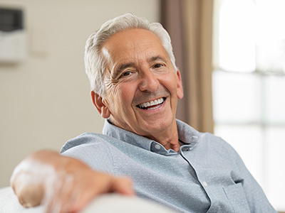 A man in a gray shirt is sitting on a couch with his arm resting on the back of it, smiling and looking directly at the camera.