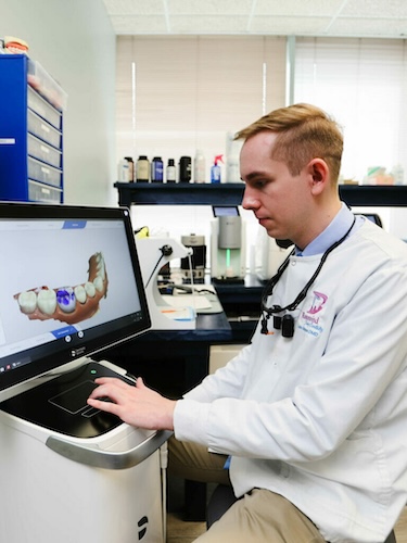 A man in a white coat is sitting at a computer monitor, looking at a 3D model of a human mouth.