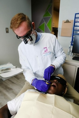 The image shows a dentist wearing protective eyewear, gloves, and a face mask, performing dental work on a patient who is lying in a chair with their eyes covered.