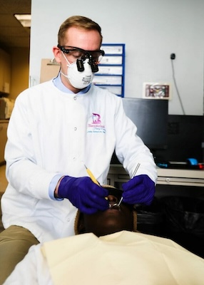 The image shows a person in a lab coat and face mask, performing what appears to be dental work on a man lying on a dental chair.