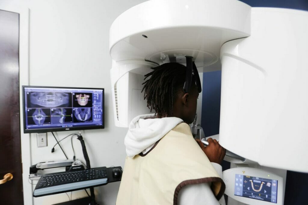 A person is seated in a medical imaging machine, likely an MRI or CT scanner, with their head inside the cylindrical opening, while another individual is standing nearby observing the scan.