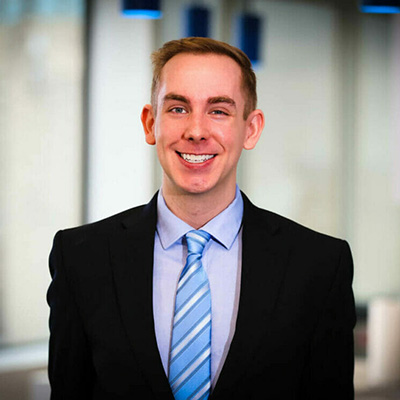 The image depicts a man standing in an office environment, wearing a suit and tie with a light blue shirt. He has short hair and is smiling at the camera.