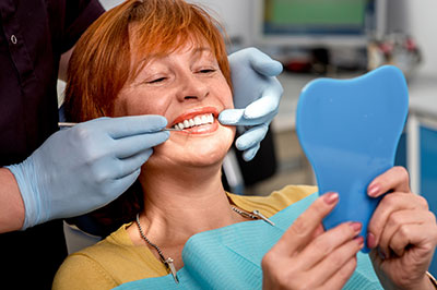 A woman wearing a blue surgical mask and holding a dental impression tray is smiling while looking at the tray, with a dentist in background.