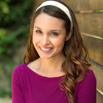 The image shows a woman with long hair and a smile, wearing a purple top, standing against a wooden fence.