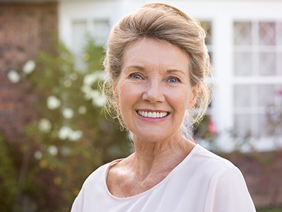 A smiling woman in a white blouse, standing outdoors in front of a brick house with a garden.