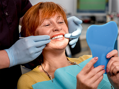 A woman in a dental chair receiving a teeth cleaning, with a dentist holding a blue model of a tooth and a mirror, both wearing gloves and masks.