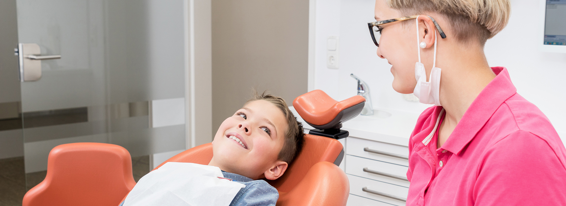 A woman in a pink shirt is assisting a young boy with dental treatment, while another person observes.