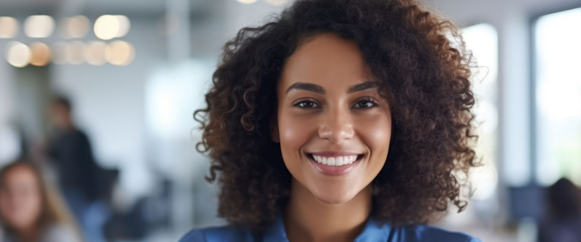 The image features a smiling woman with curly hair, wearing a blue top, standing in an office environment.