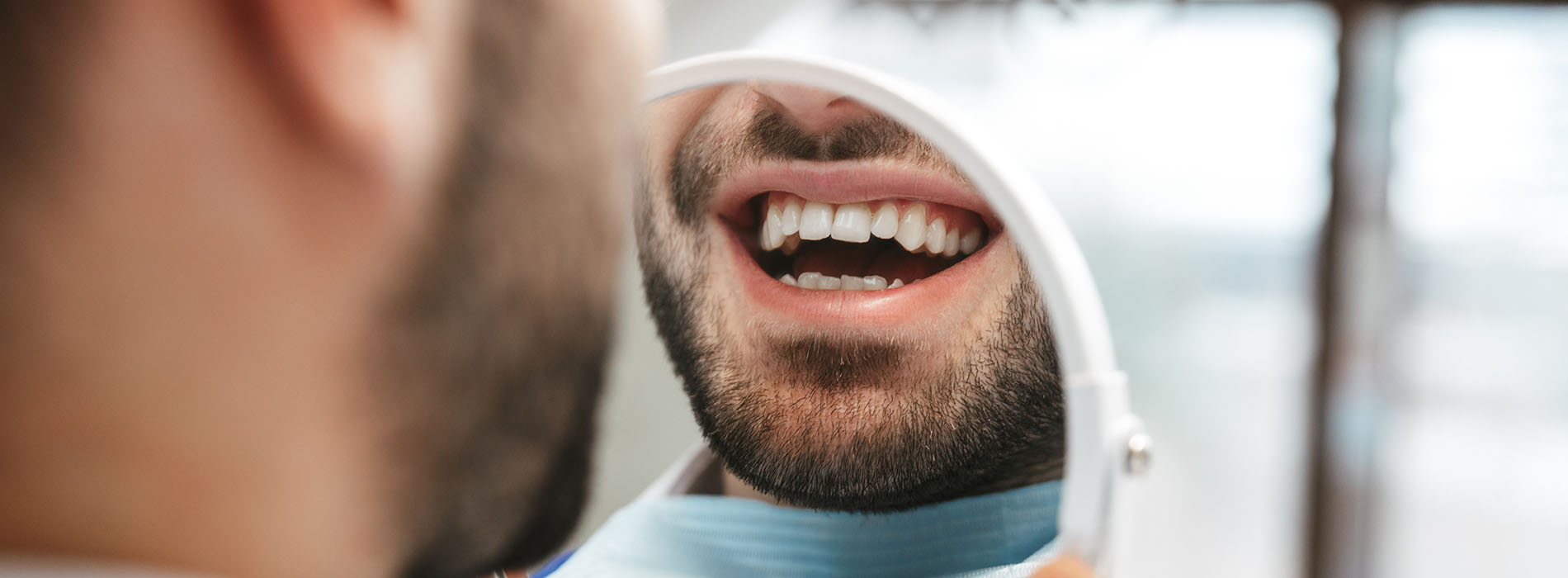 The image shows a close-up of a man with his mouth open, displaying white teeth and the interior of his mouth, with a blurred background that appears to be an indoor setting.