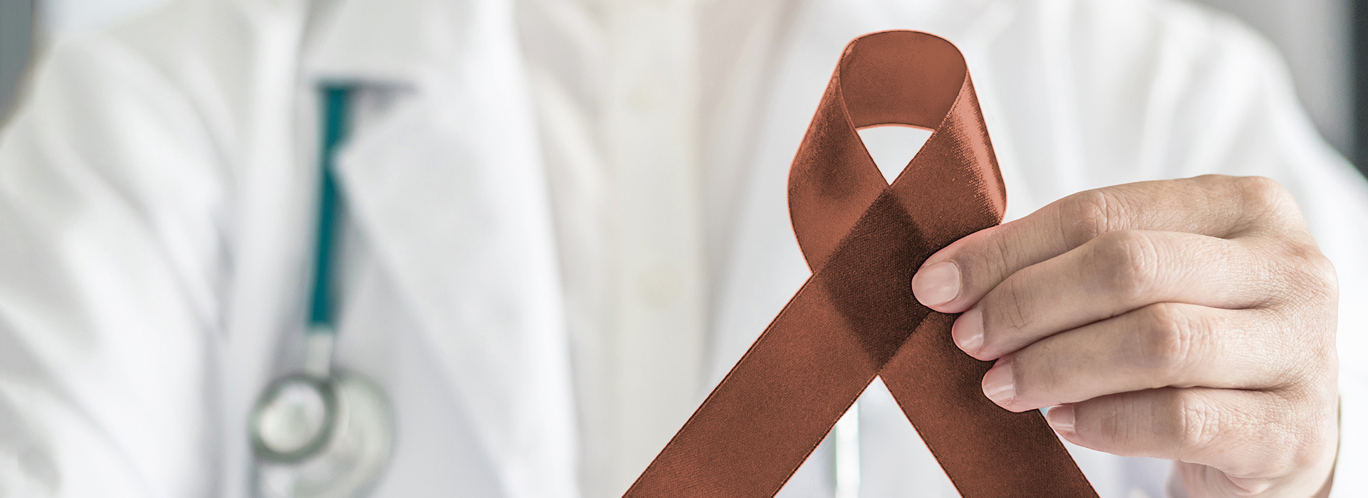 The image is a photograph of a person holding a brown ribbon with a red symbol on it, which appears to be related to medical awareness or support. The individual is wearing a white coat and stethoscope, suggesting they are a healthcare professional, possibly a doctor or nurse.