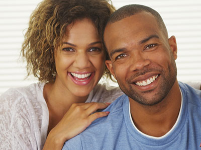 The image depicts a man and woman smiling at the camera, with the man wearing a short beard and the woman having curly hair. They appear to be in a happy mood, possibly posing for a portrait or a casual photograph.