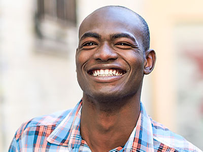 The image is a photograph of a smiling man with short hair, wearing a plaid shirt and standing outdoors.