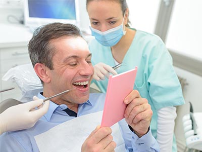 A man sitting in a dental chair, smiling at a pink card he s holding, while a dental professional stands behind him with a tray of instruments and a surgical mask.