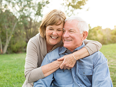 An elderly couple embracing in a park, with the man wearing a blue shirt and the woman in a brown jacket.