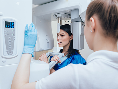 A woman is standing in front of a 3D imaging machine, possibly a dental or medical scanner, with a healthcare professional examining the screen behind her.