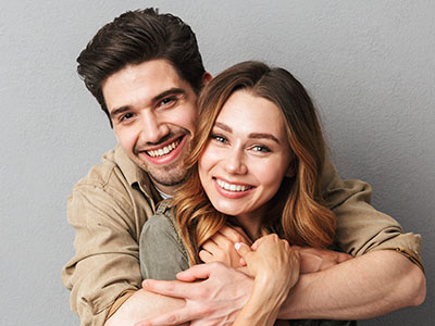 A young couple embracing and smiling, with the man wearing a light brown shirt and the woman in a dark top.