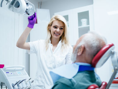 A woman in a white lab coat is assisting an elderly man with a medical device while he is seated in a dental chair.