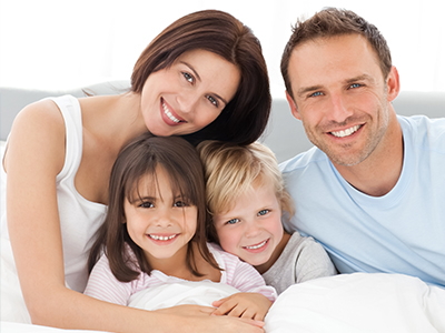 A family of four, including two adults and two children, posing for a photograph in bed with smiles on their faces.