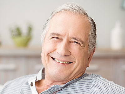 The image shows a smiling older man with gray hair, wearing a blue shirt and sitting comfortably in a chair at home.