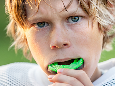 The image shows a young male with blond hair and blue eyes, wearing a football uniform, holding a green toothbrush to his mouth.