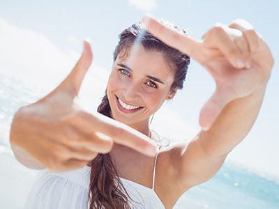 A woman is smiling and holding up a camera to take a selfie, with the ocean in the background.